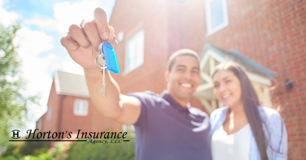 A close-up of a hand holding up a house key on a blue keychain with a smiling couple and a new brick home blurred in the bright background